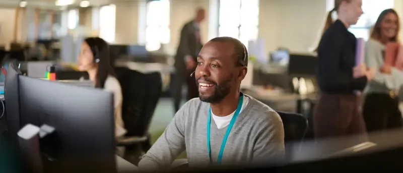 A smiling male call center employee wearing a headset looks at his computer screen in a busy open-plan office.